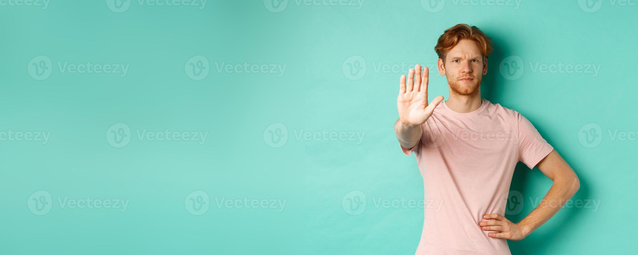 Serious young man with red hair and beard telling to stop, showing palm and frowning, disapprove and prohibit something bad, standing over turquoise background photo