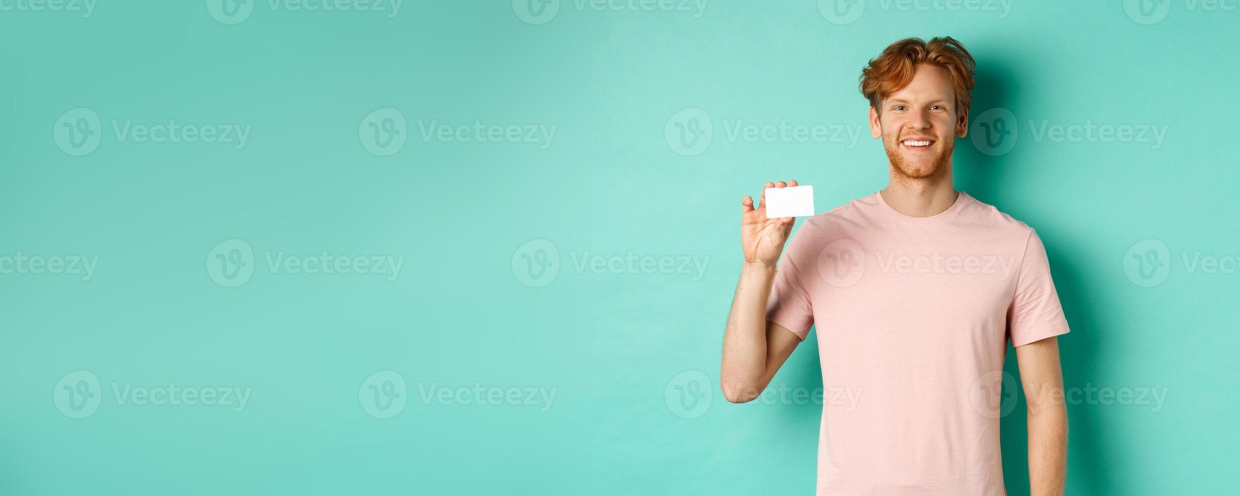 Handsome young man smiling and showing plastic credit card, standing in t-shirt against turquoise background photo