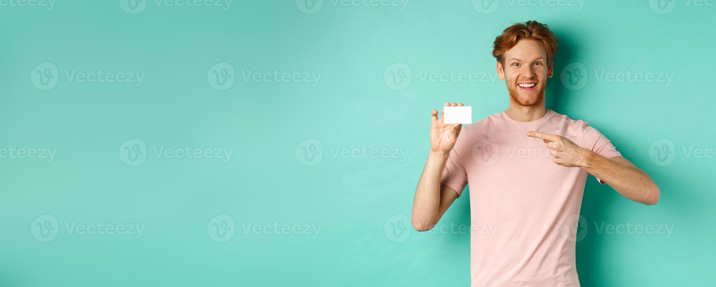Attractive adult man with beard and red hair pointing finger at plastic credit card, smiling pleased at camera, standing over turquoise background photo