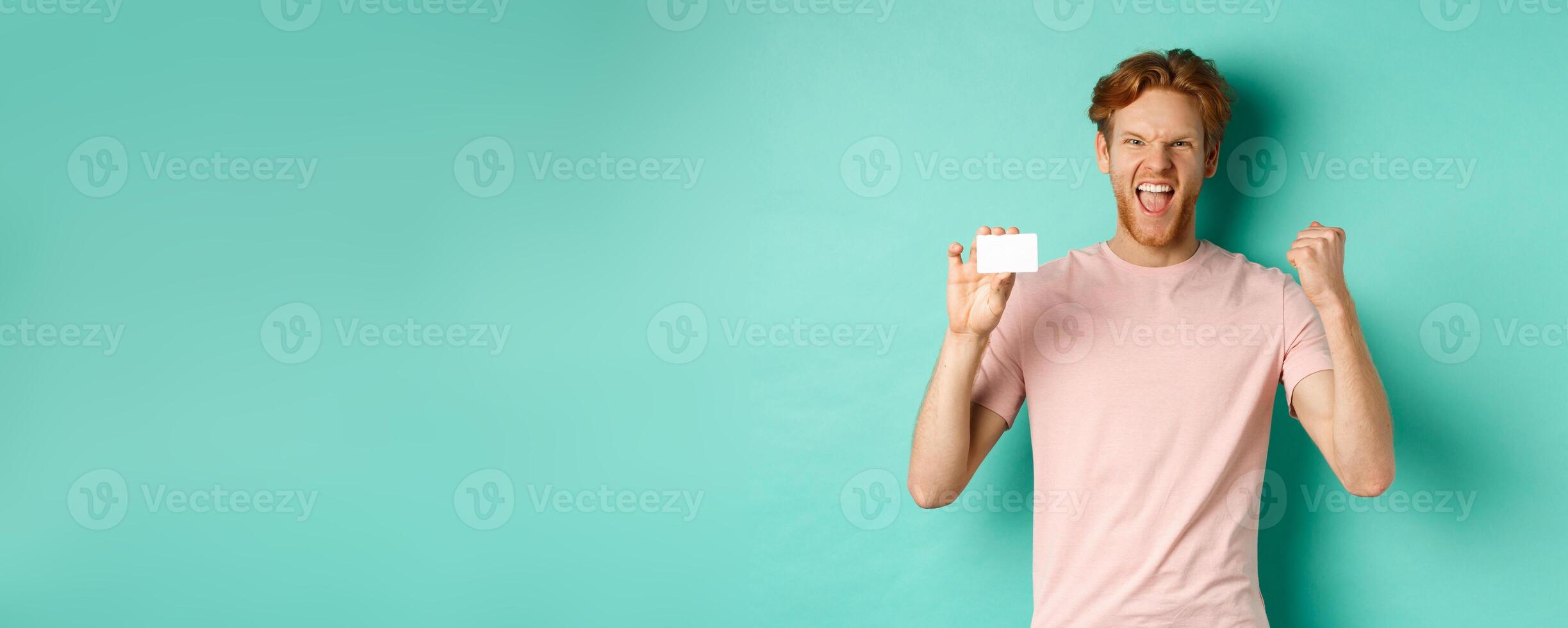Cheerful young man triumphing, making fist pump to celebrate success, showing plastic credit card, winning prize from bank, turquoise background photo