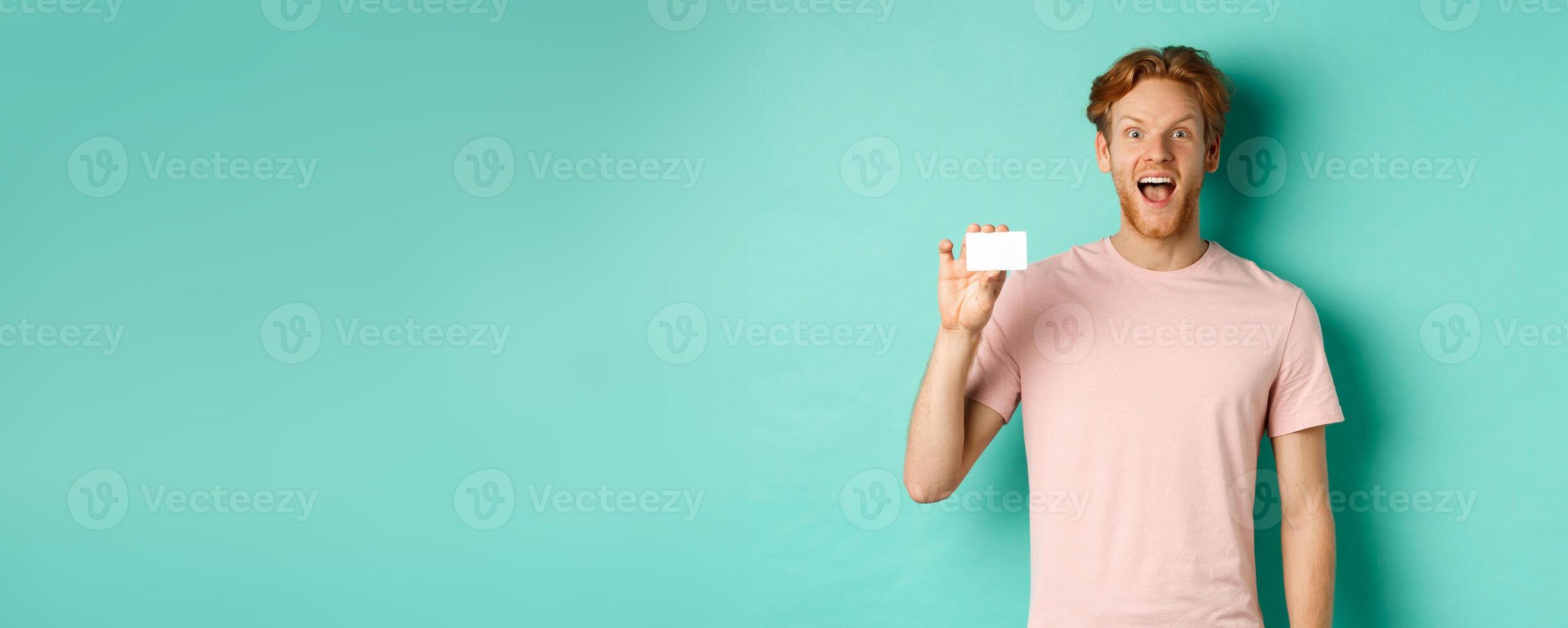 Cheerful young man with red hair and beard, wearing t-shirt, showing plastic credit card and smiling at camera, demonstrate new bank promo photo
