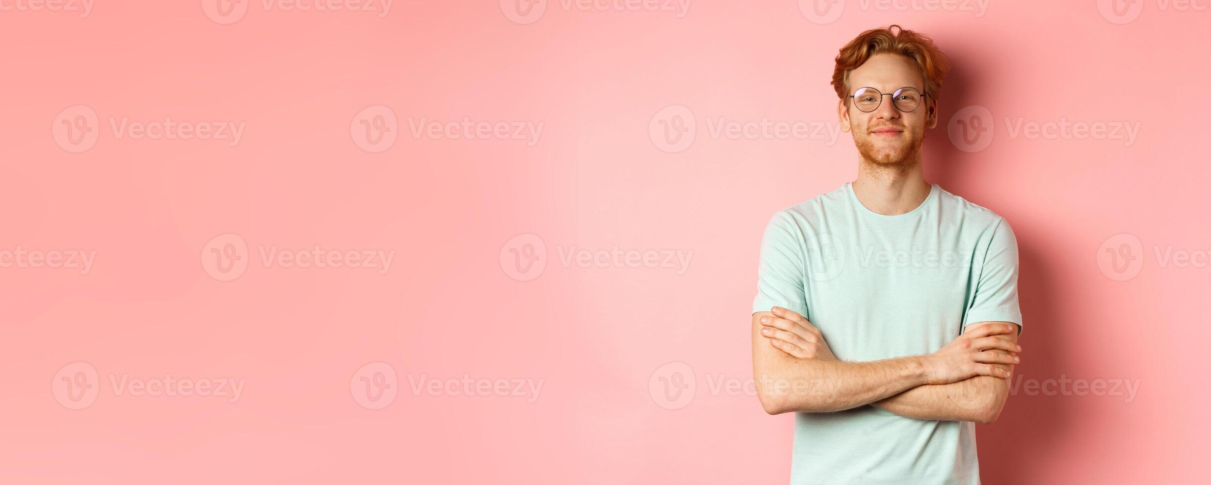 Portrait of satisfied caucasian man with red hair and beard, cross arms on chest and smiling with smug face, wearing glasses, standing over pink background photo