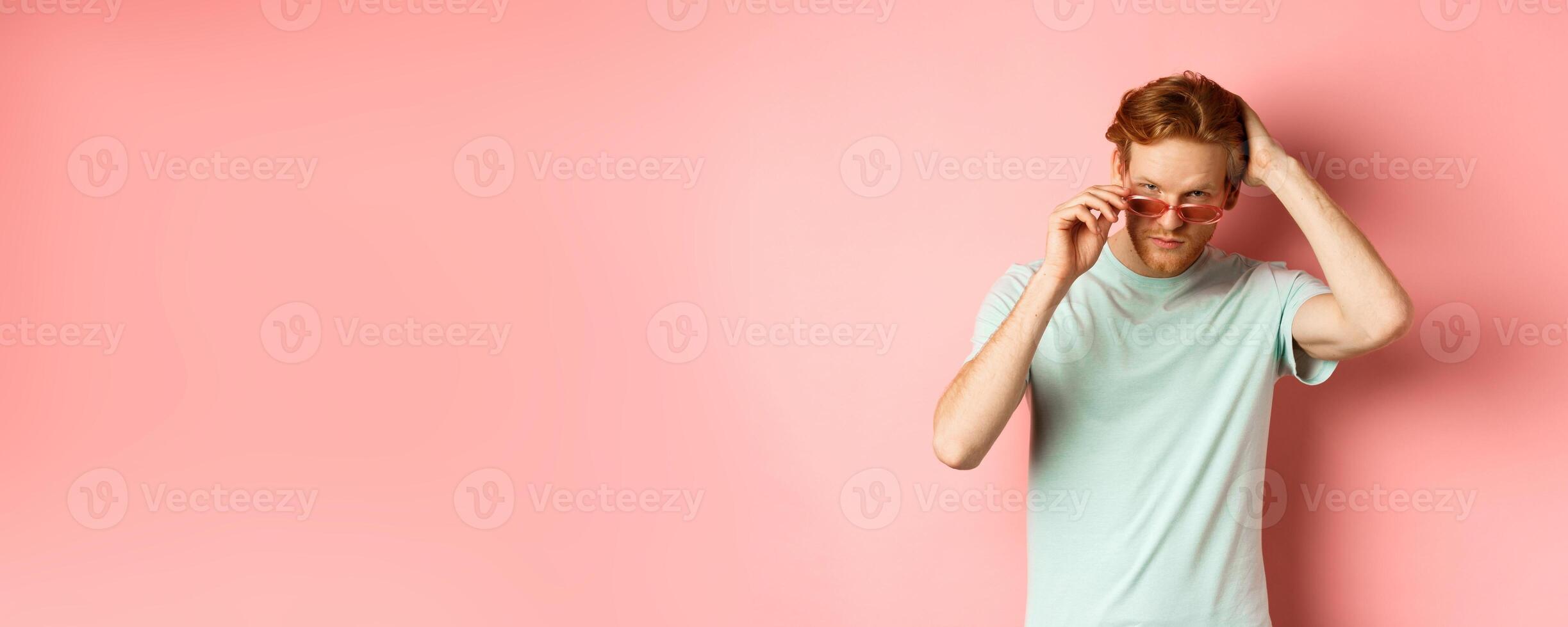 Handsome young redhead man in sunglasses, brushing hair with hand and looking smug and confident at camera, standing over pink background photo
