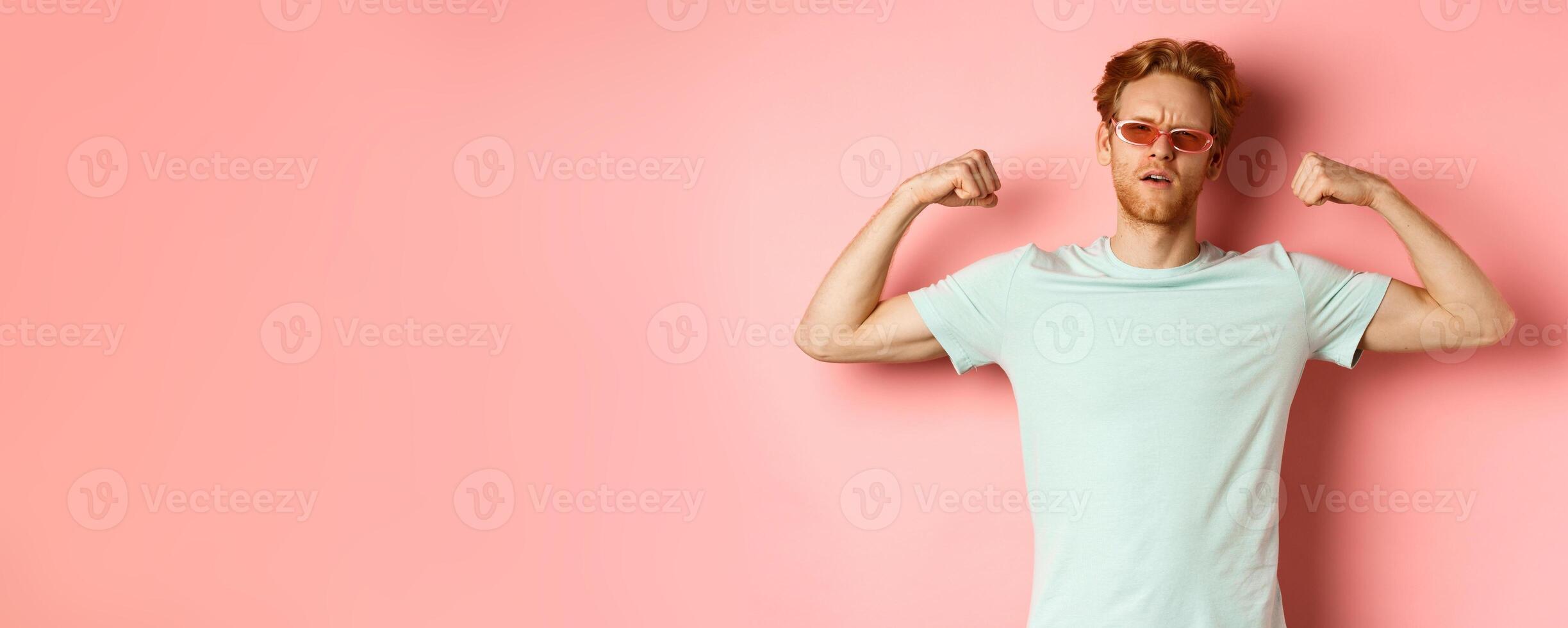 Confident young man with red hair, wearing summer sunglasses and t-shirt, showing strong and fit body muscles, flex biceps and staring cool at camera, pink background photo