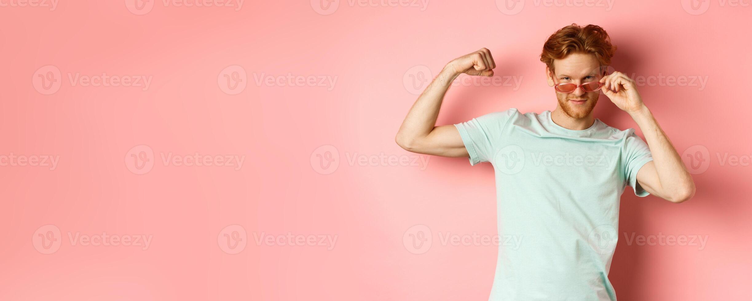 Confident young man with red hair, wearing summer sunglasses and t-shirt, showing strong and fit body muscles, flex biceps and staring cool at camera, pink background photo