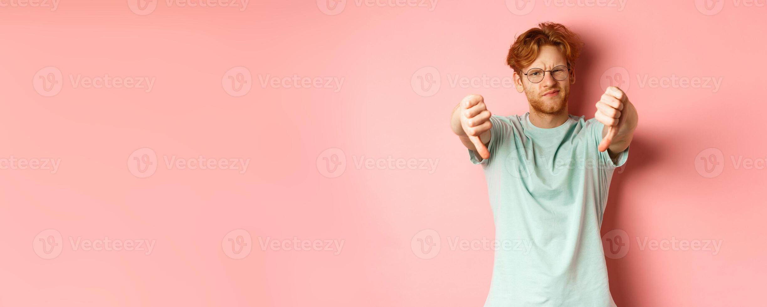Disappointed young man in glasses, with messy red haircut, showing thumbs down and grimacing unsatisfied, express dislike, standing over pink background photo
