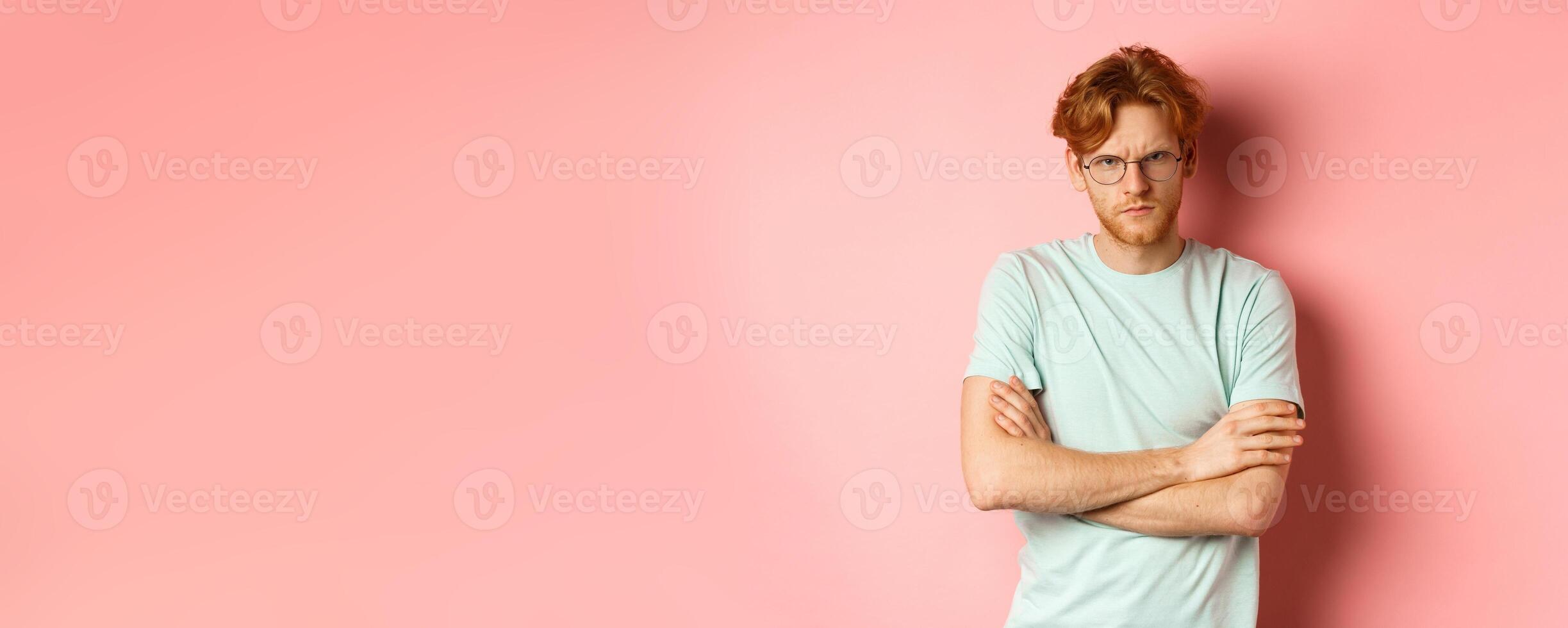 Angry man in glasses with red hair frowning, cross arms on chest in defensive pose, sulking at you, standing over pink background photo