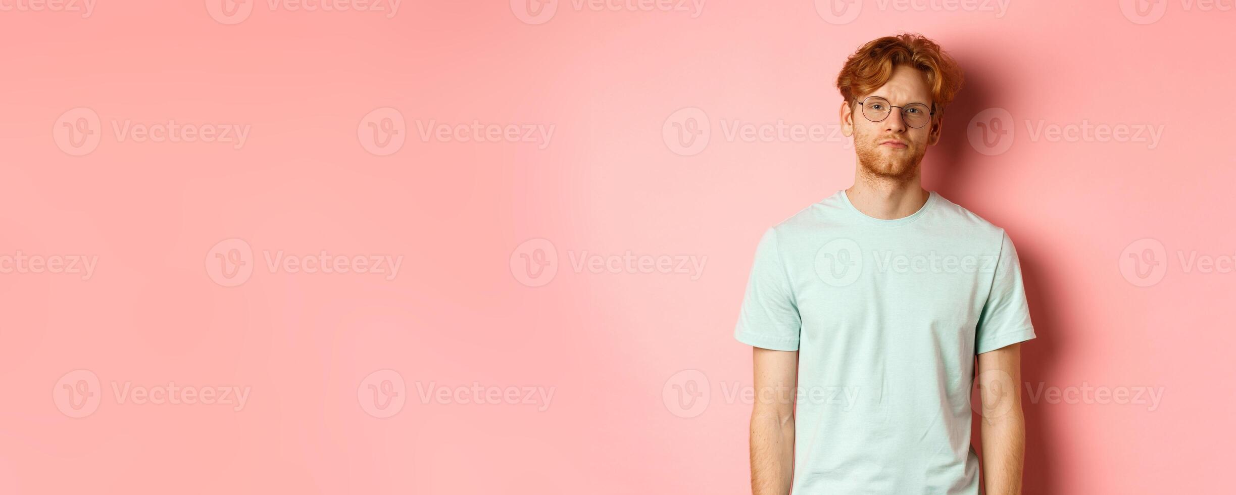 Sad and gloomy redhead bearded man in t-shirt and glasses, staring at camera bored and unamused, standing over pink background photo