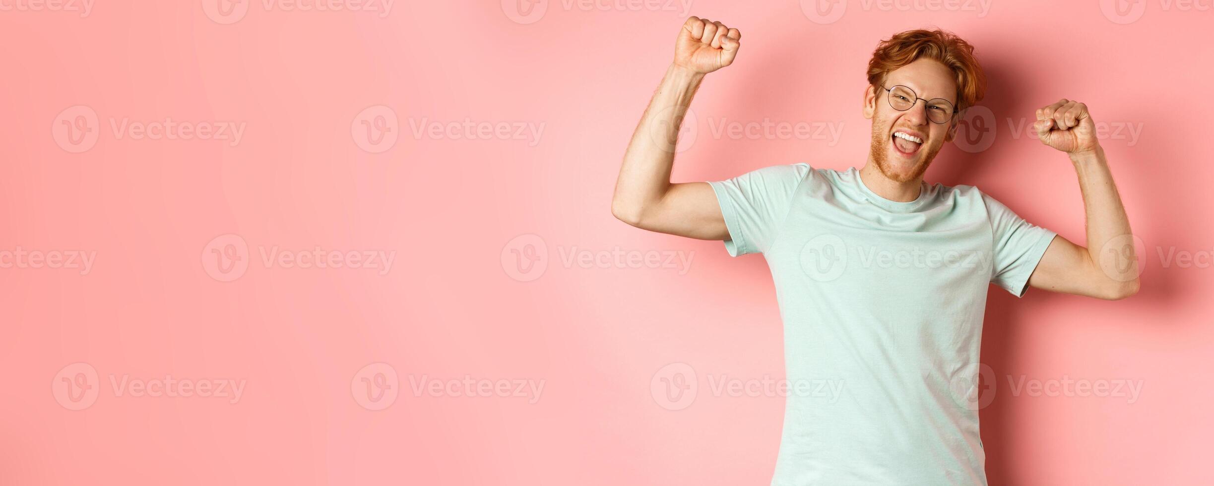 Cheerful young man with red hair looking happy, raising hands up in fist pumps gesture, celebrating success, feel like champion, winning and standing over pink background photo