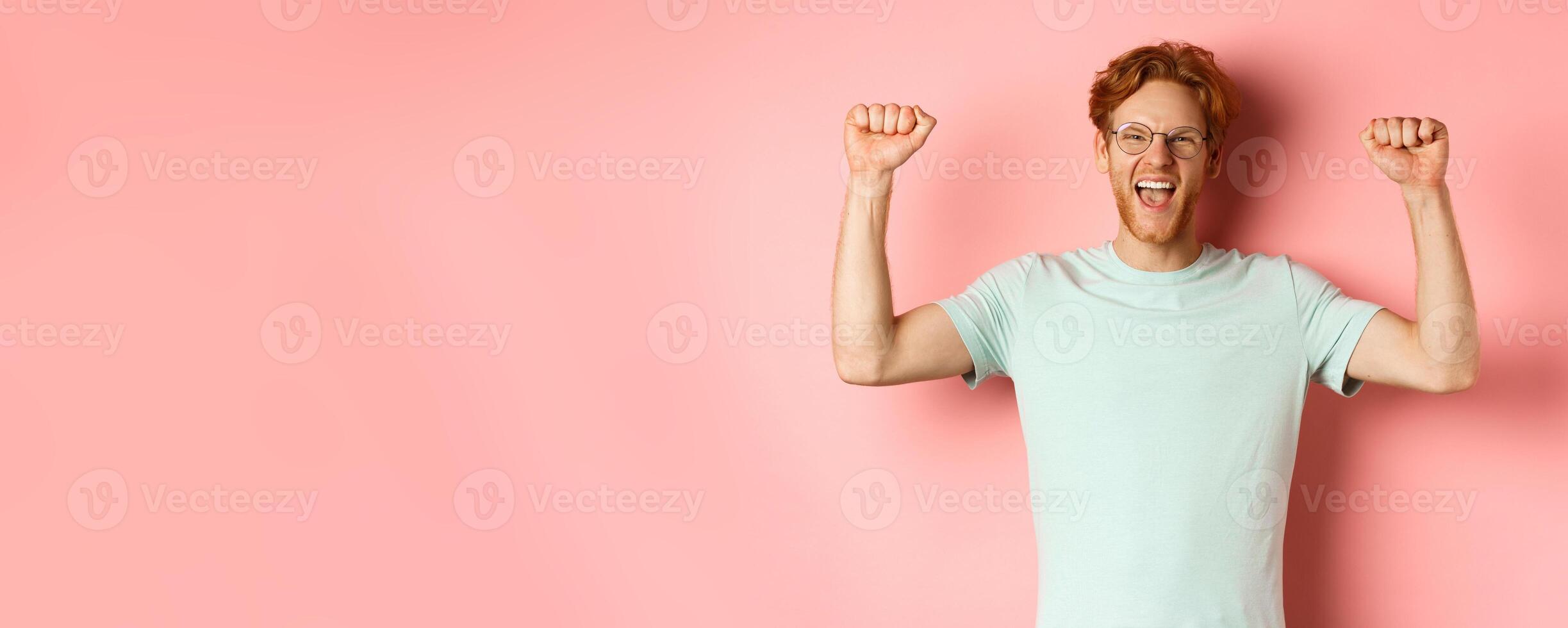 Happy winner with red hair and beard celebrating victory, shouting yes with joy and raising hands up, enjoy success, standing over pink background photo