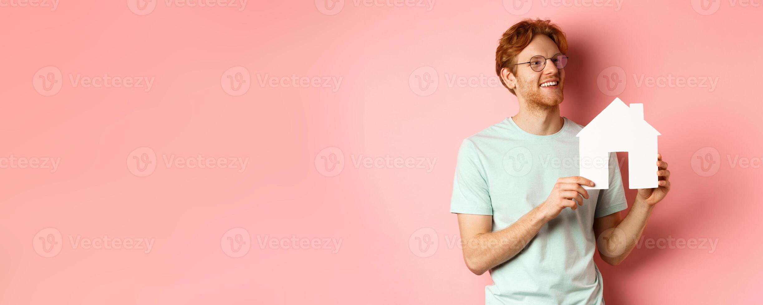 Real estate. Happy redhead man dreaming of buying property, looking right and showing paper house cutout, standing over pink background photo