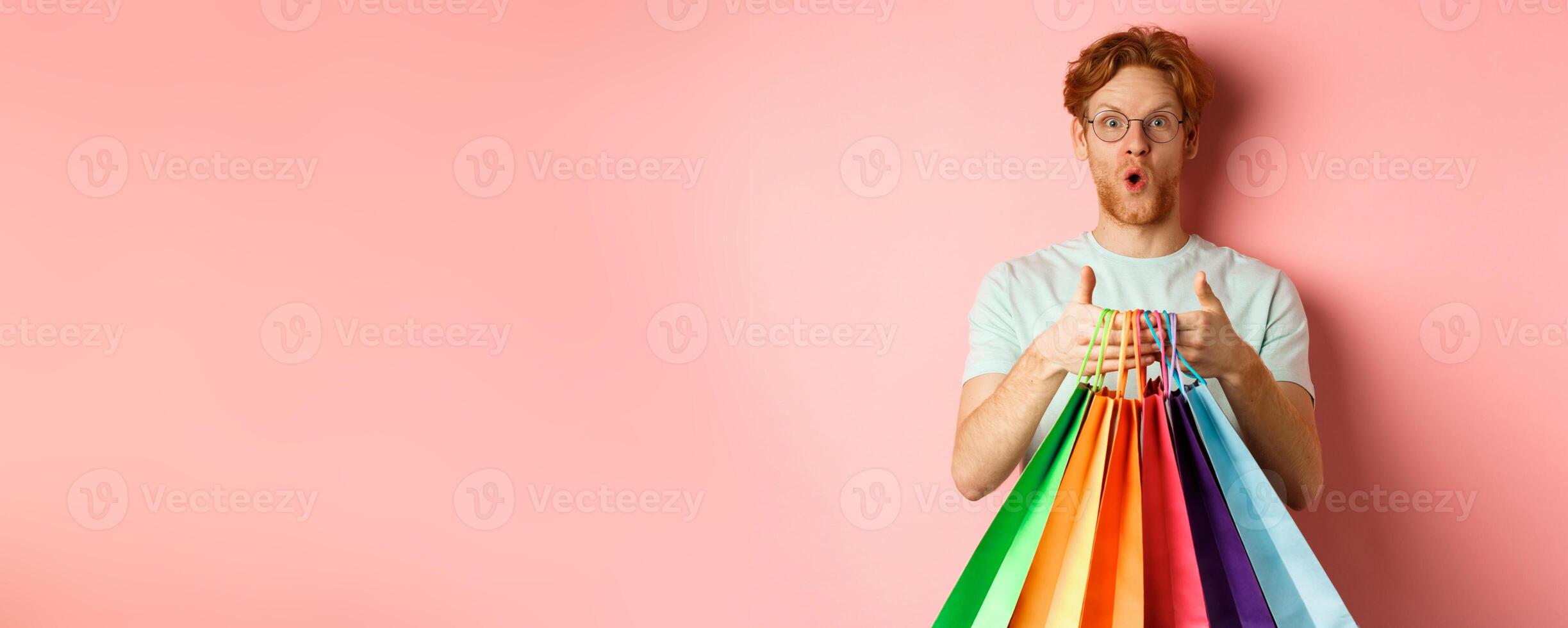 Young handsome man amazed with special discounts, buying gifts, holding shopping bags and standing over pink background photo