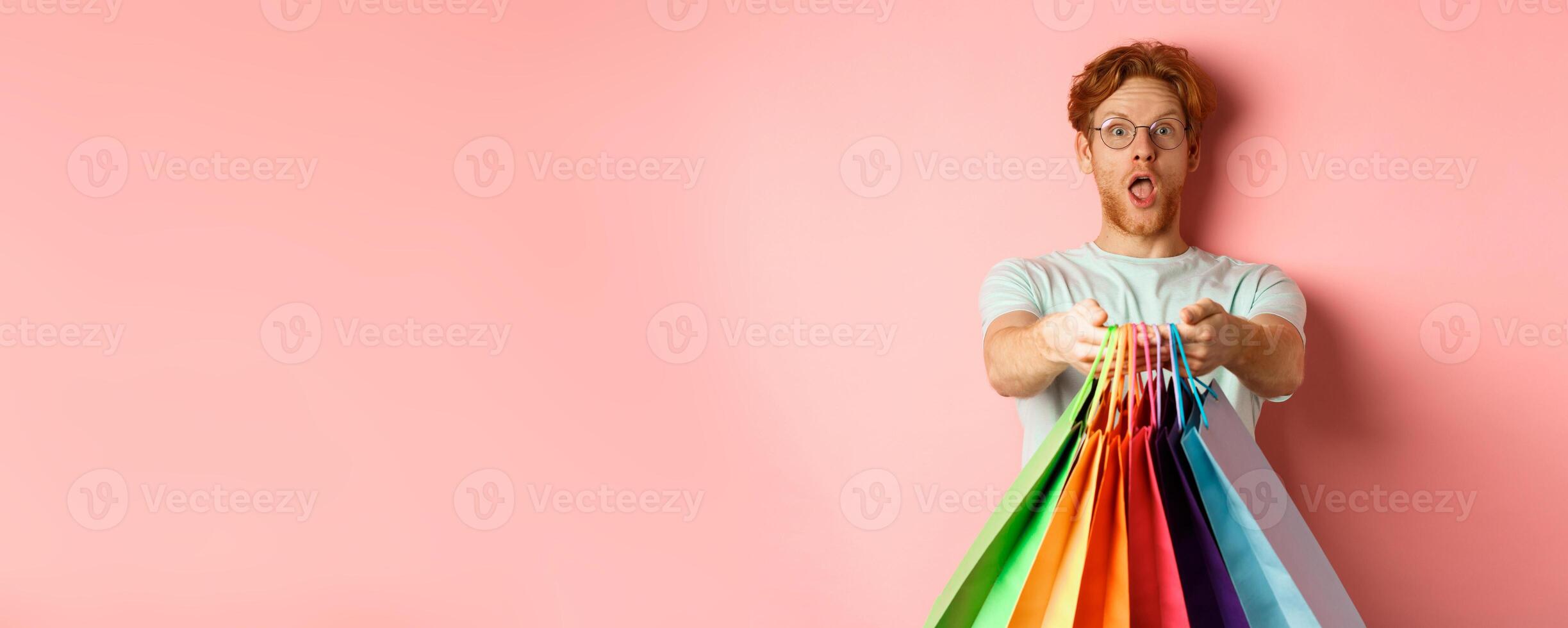Surprised redhead man stretch out hands with shopping bags, give you gifts, standing over pink background photo