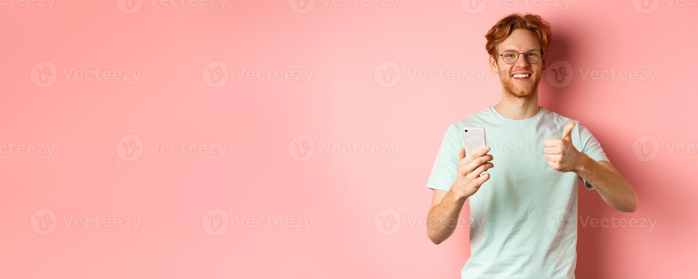 Happy young man with red messy hair, showing thumbs-up while using mobile phone, smiling and praising application, standing over pink background photo