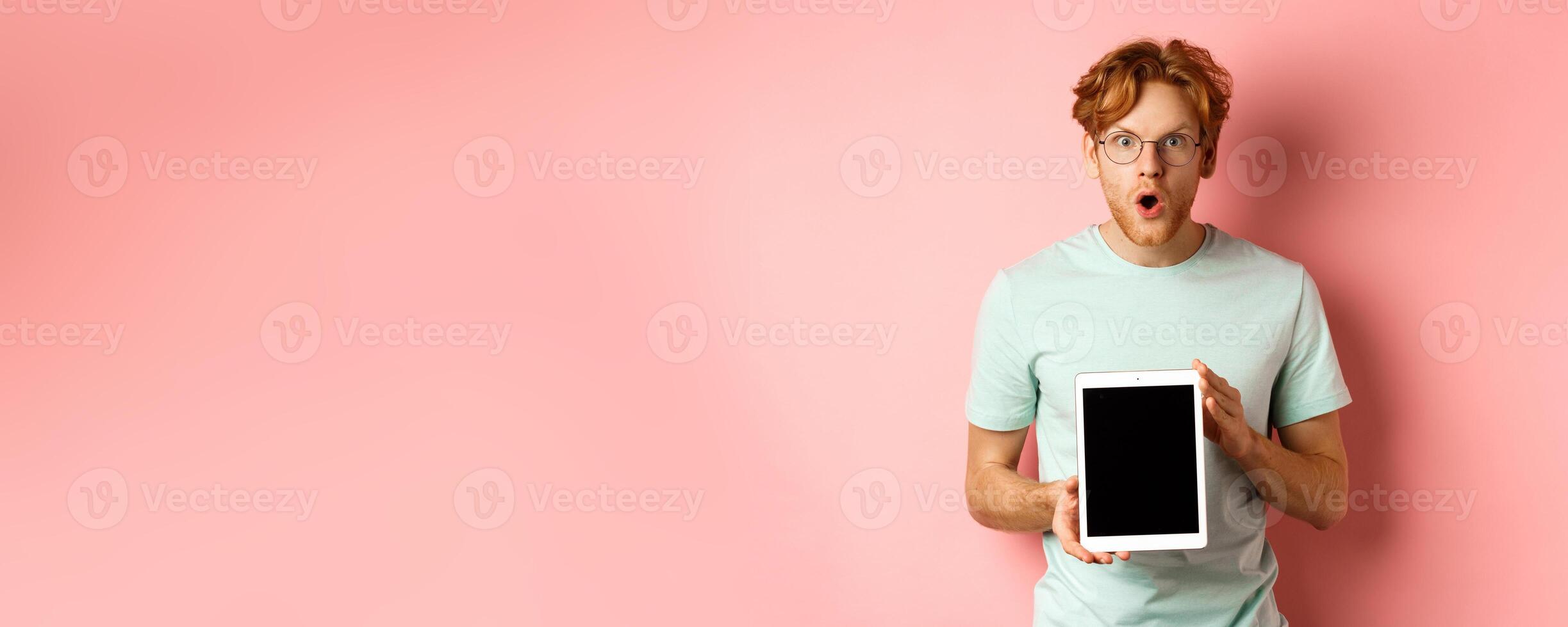 Excited young man with red hair and beard, checking out online promotion, showing digital tablet screen and staring at camera amazed, standing over pink background photo