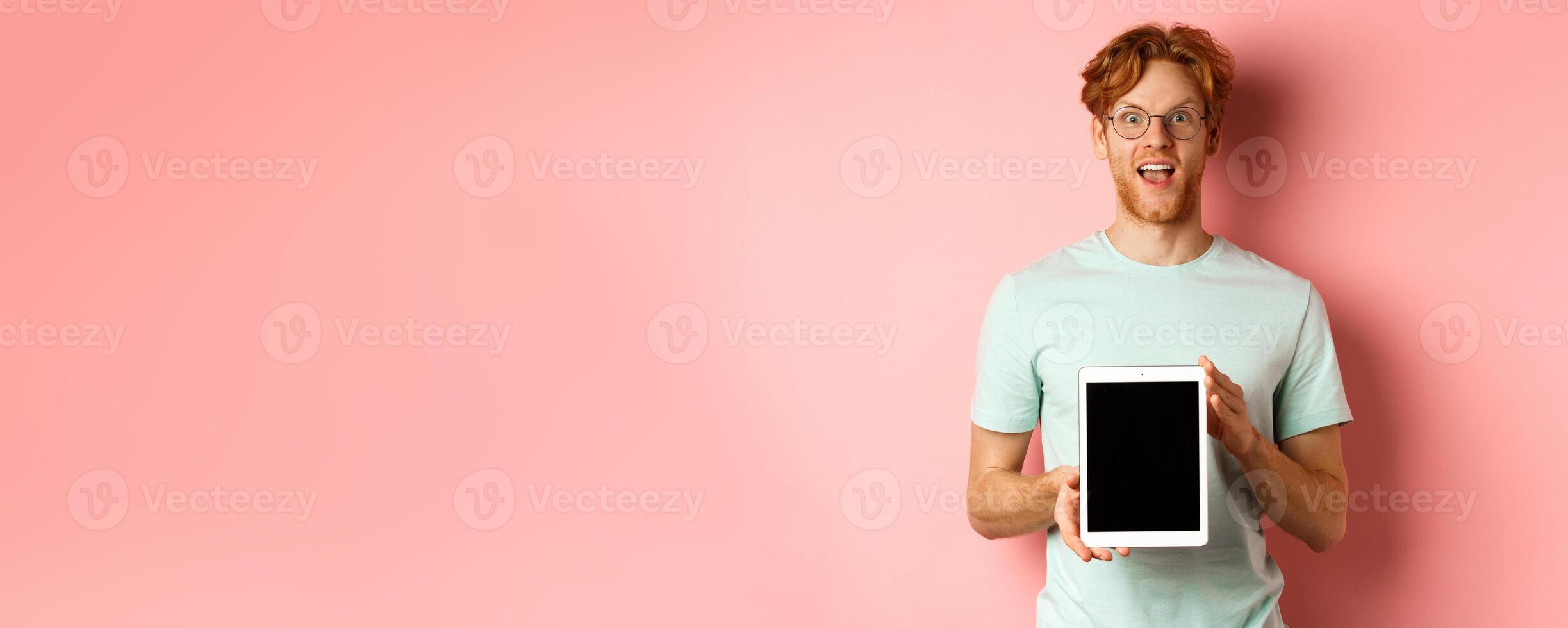 Excited young man with red hair and beard, checking out online promotion, showing digital tablet screen and staring at camera amazed, standing over pink background photo