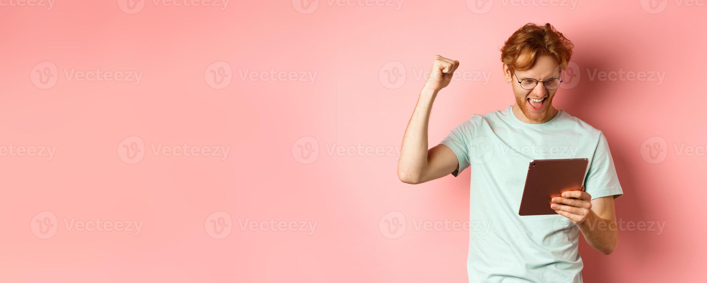 Image of happy redhead man triumphing, winning online with digital tablet and rejoicing, standing over pink background photo