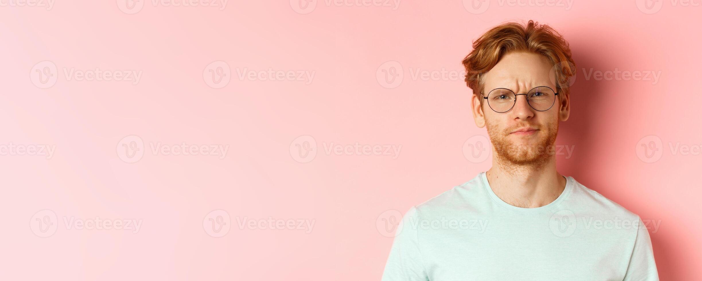 Headshot of skeptical redhead man in glasses and t-shirt frowning disappointed, staring with disapproval and judgement, standing over pink background photo