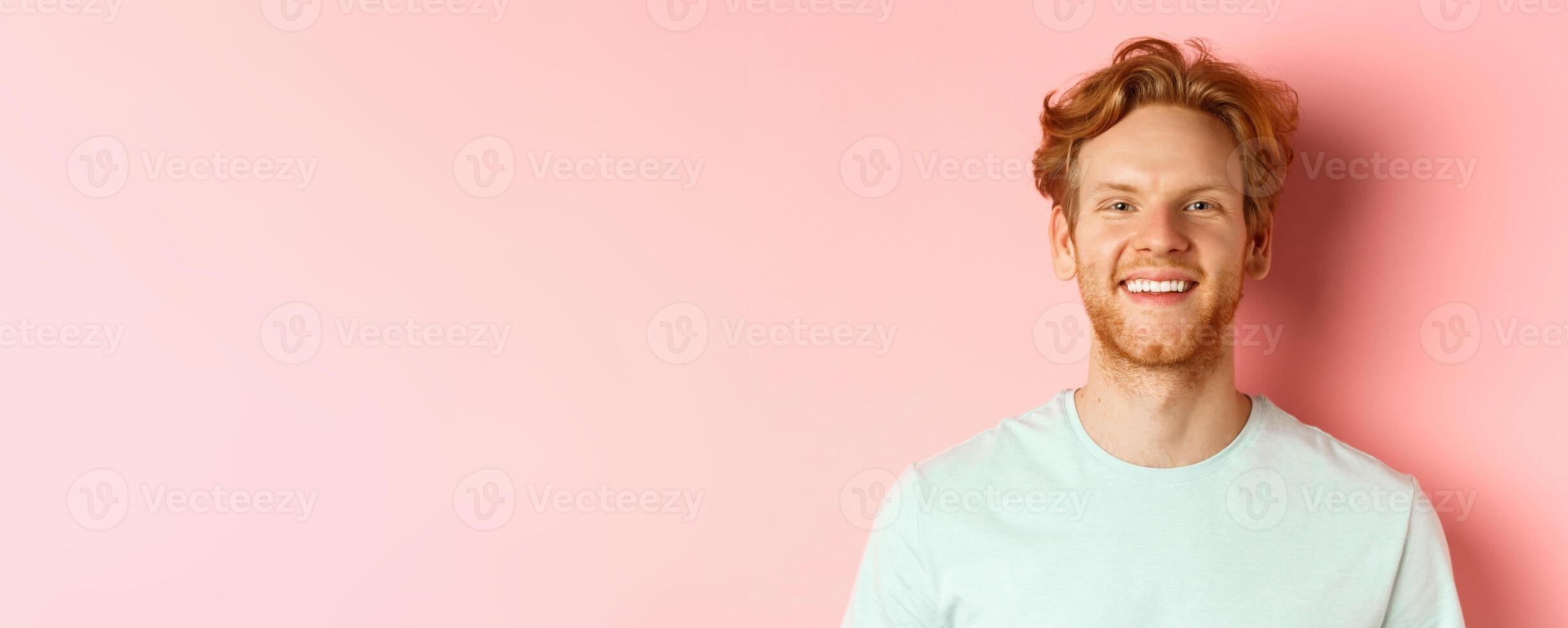 Happy young man with beard and messsy red haircut, smiling with white teeth and cheerful expression, standing over pink background photo