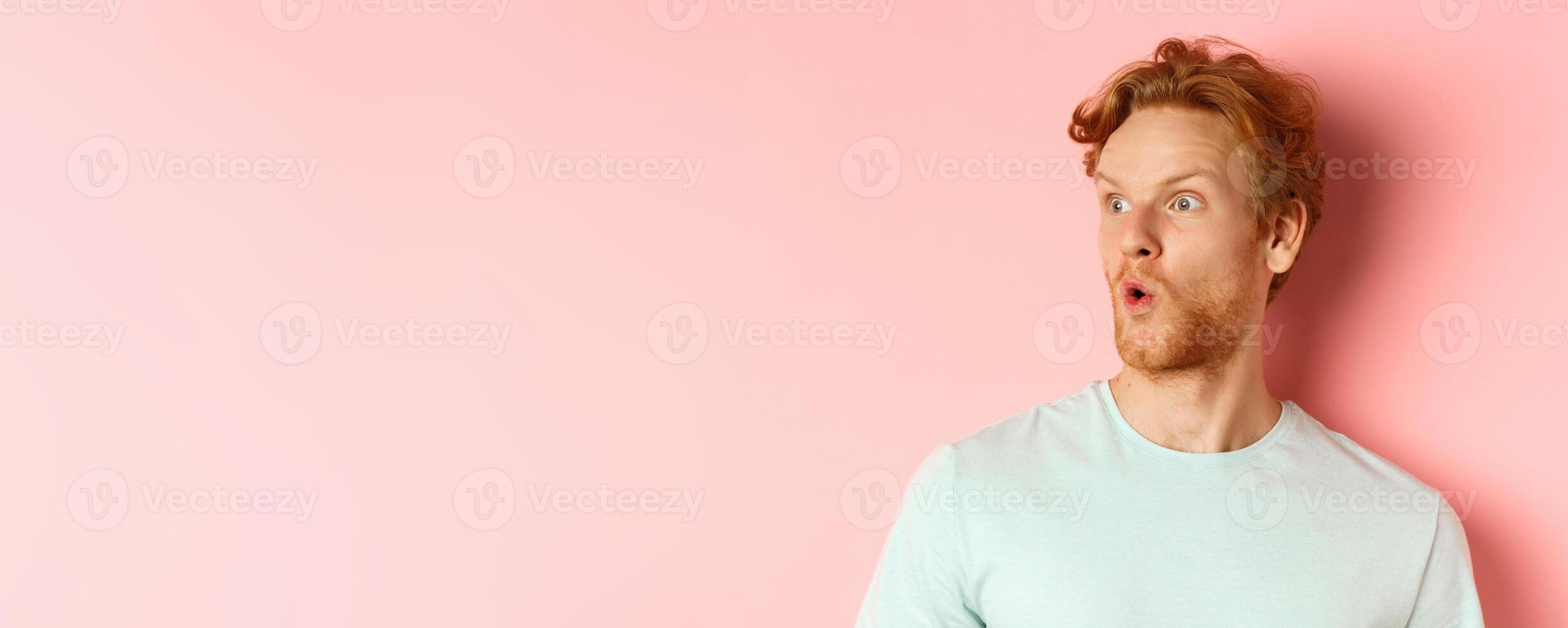 Headshot portrait of surprised redhead man with beard, looking left and saying wow, raising eyebrows amazed, standing over pink background photo