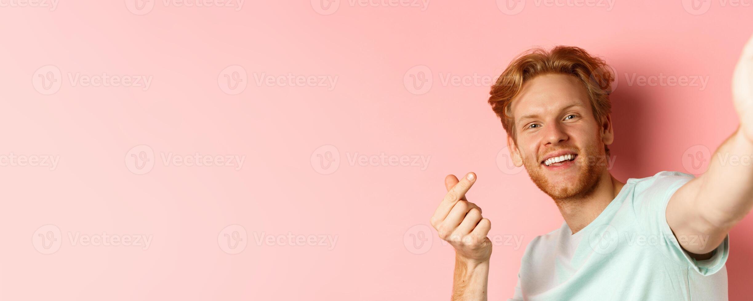 Handsome young man with beard and red hair taking selfie against pink background, stretch out hand to hold smartphone camera and showing korean heart gesture, pink background photo