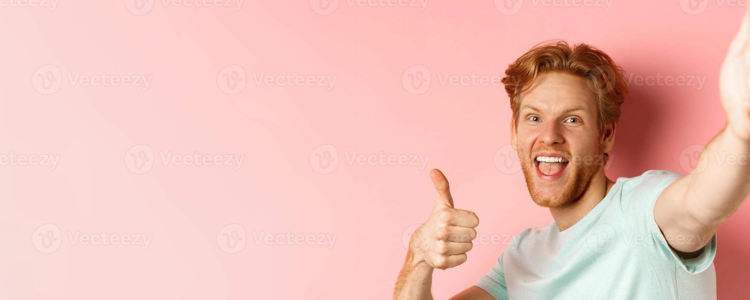 Excited redhead man tourist taking selfie and showing thumbs-up, holding camera with stretched out hand, standing over pink background photo