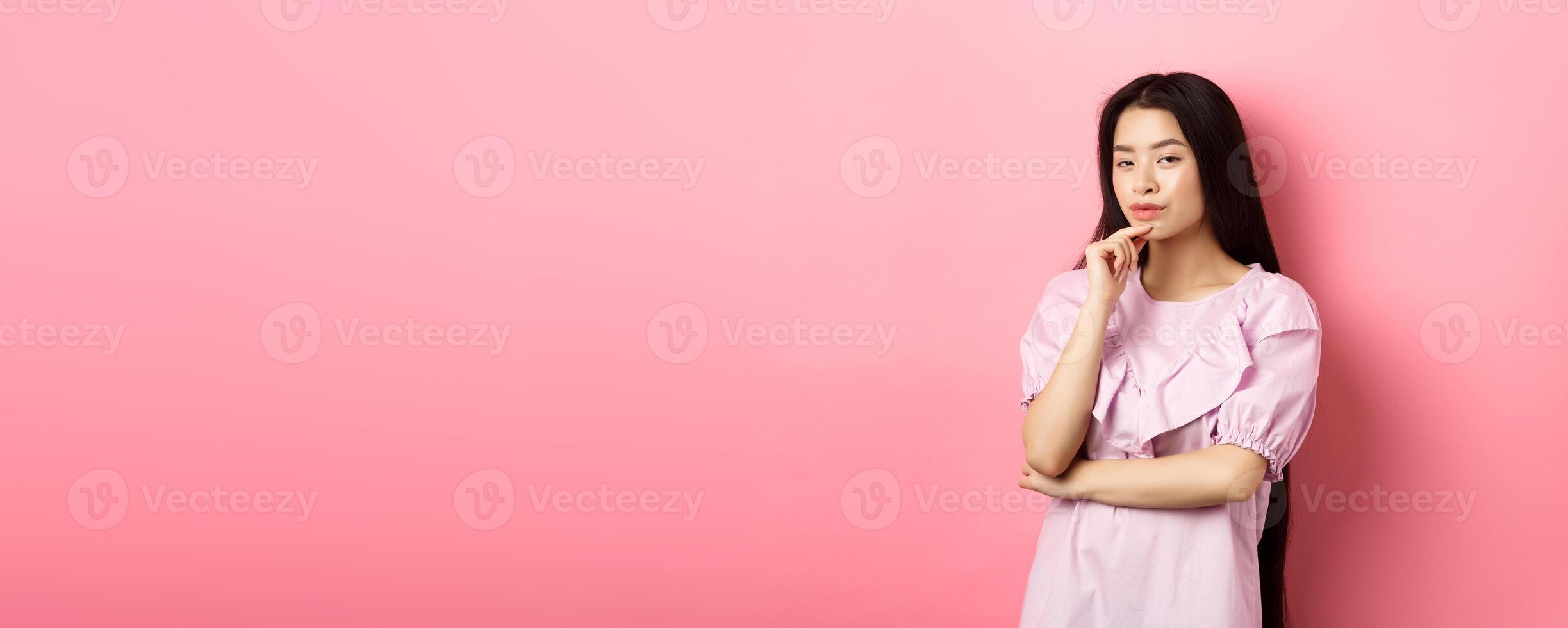 Hmm, interesting. Pensive and cunning asian girl having idea, plan to do something, looking at camera and touching chin with thoughtful face, standing on pink background photo