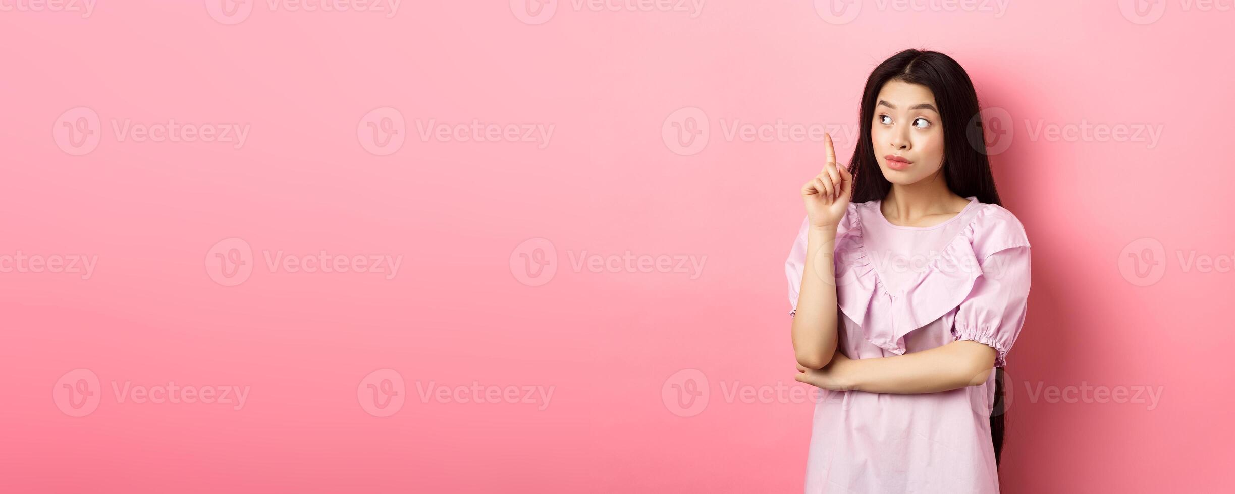 Beautiful asian girl raising finger and looking left at logo, having an idea, found solution, standing against pink background photo