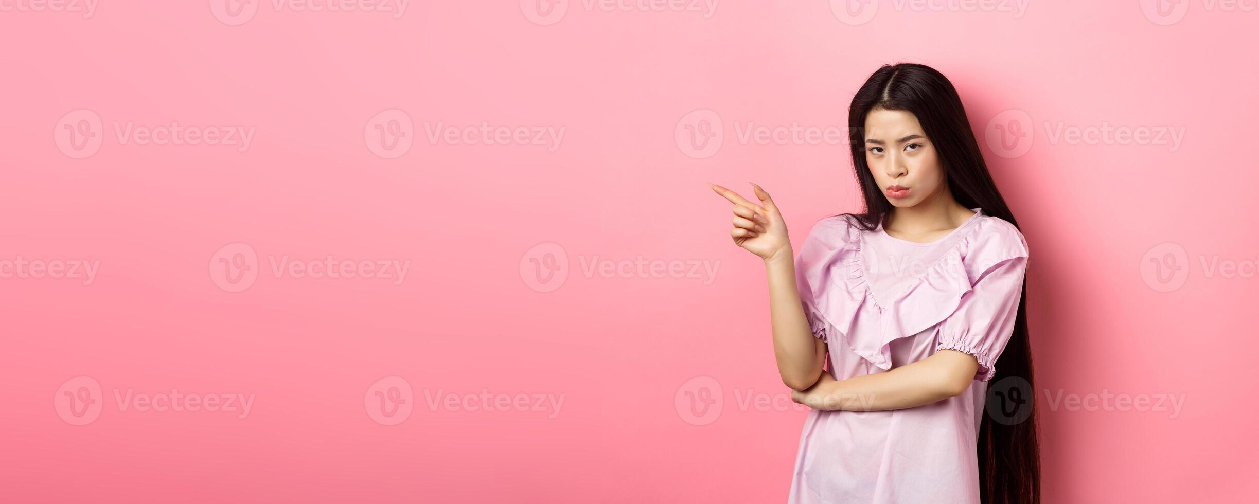 Sad and grumpy asian girl sulking, pointing left and look at camera jealous, want something, complaining and showing desired thing, standing on pink background photo
