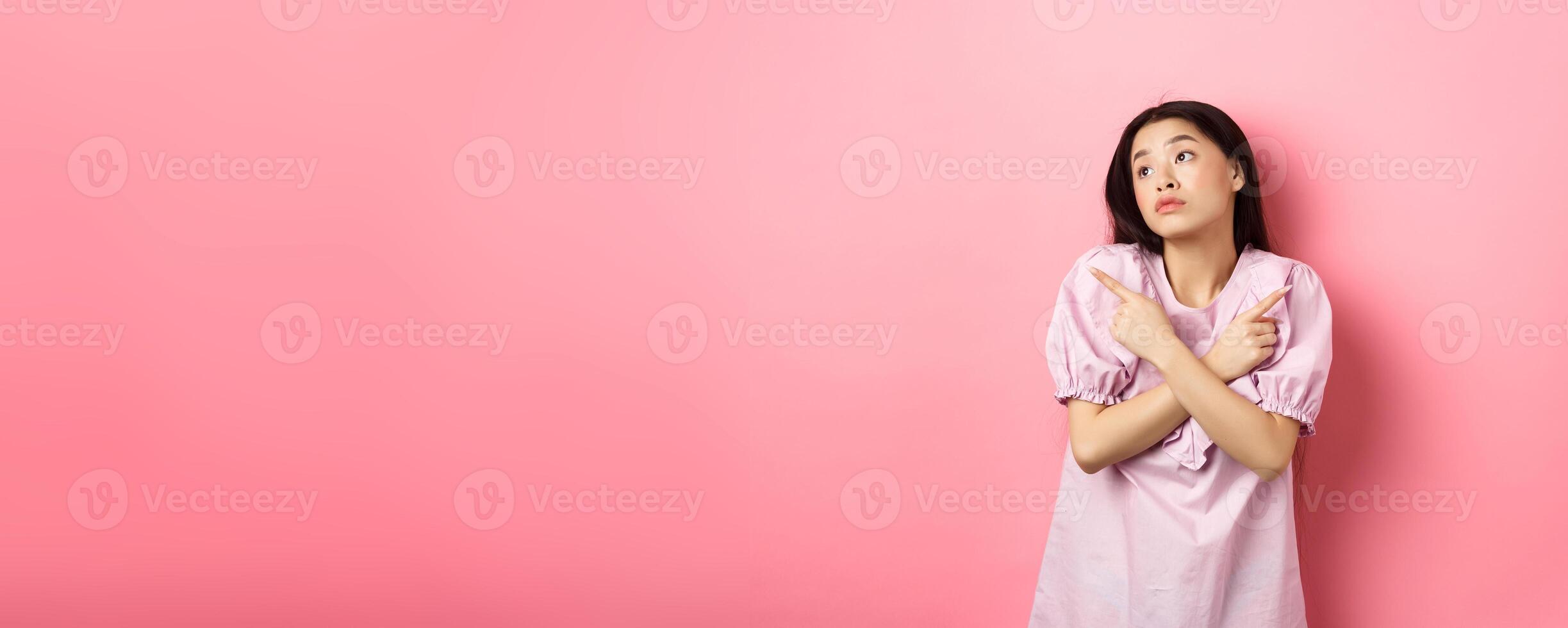 Indecisive asian woman cant decide, pointing fingers sideways and look pensive aside, making choice, standing confused on pink background photo