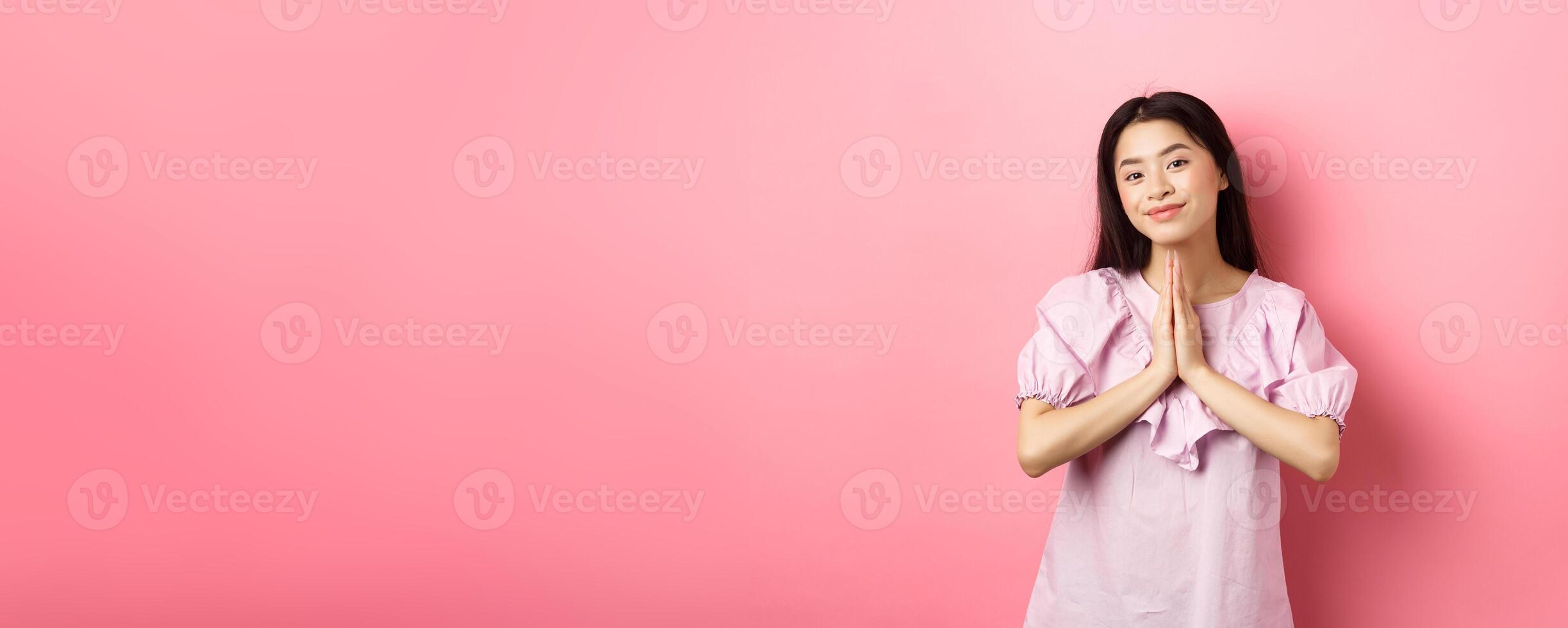 Cute asian girl say thank you, smiling and looking happy, showing namaste gesture in gratitude, standing in dress against pink background photo
