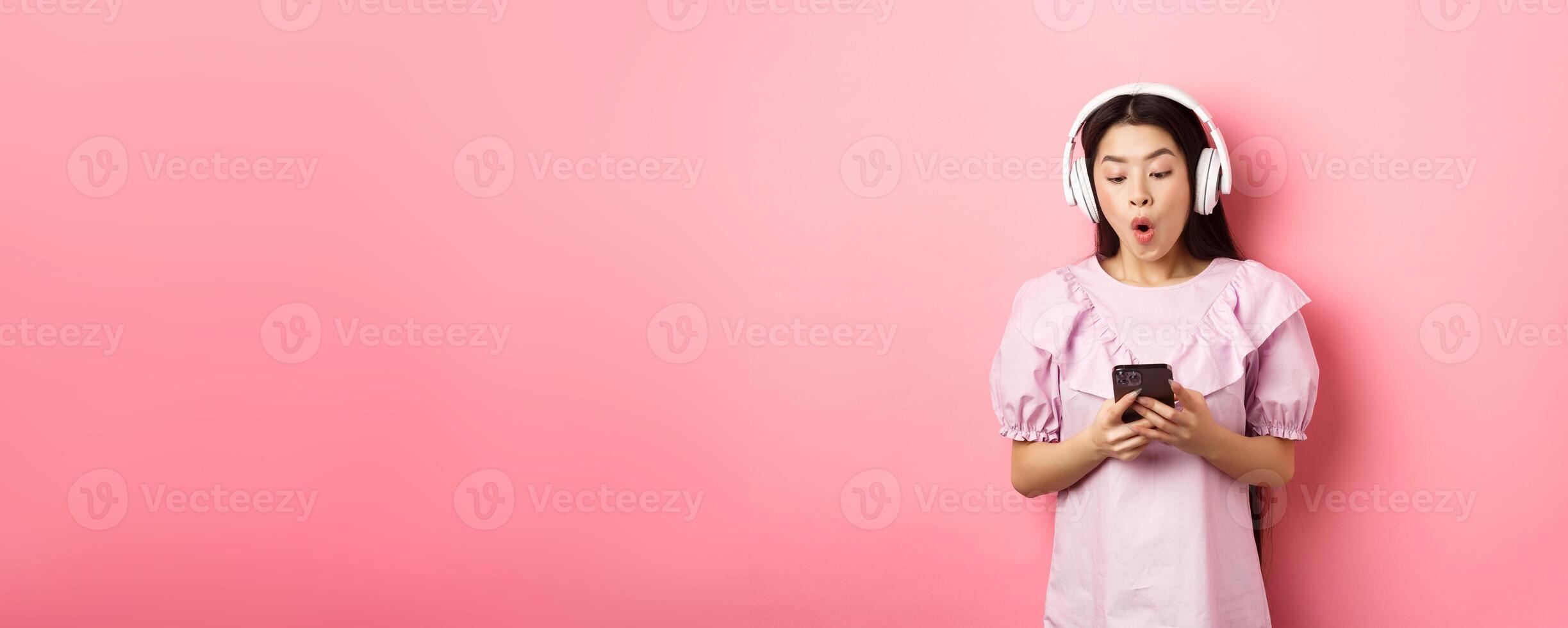Excited asian girl looking at smartphone screen amused, say wow, listening music in wireless headphones, standing against pink background photo