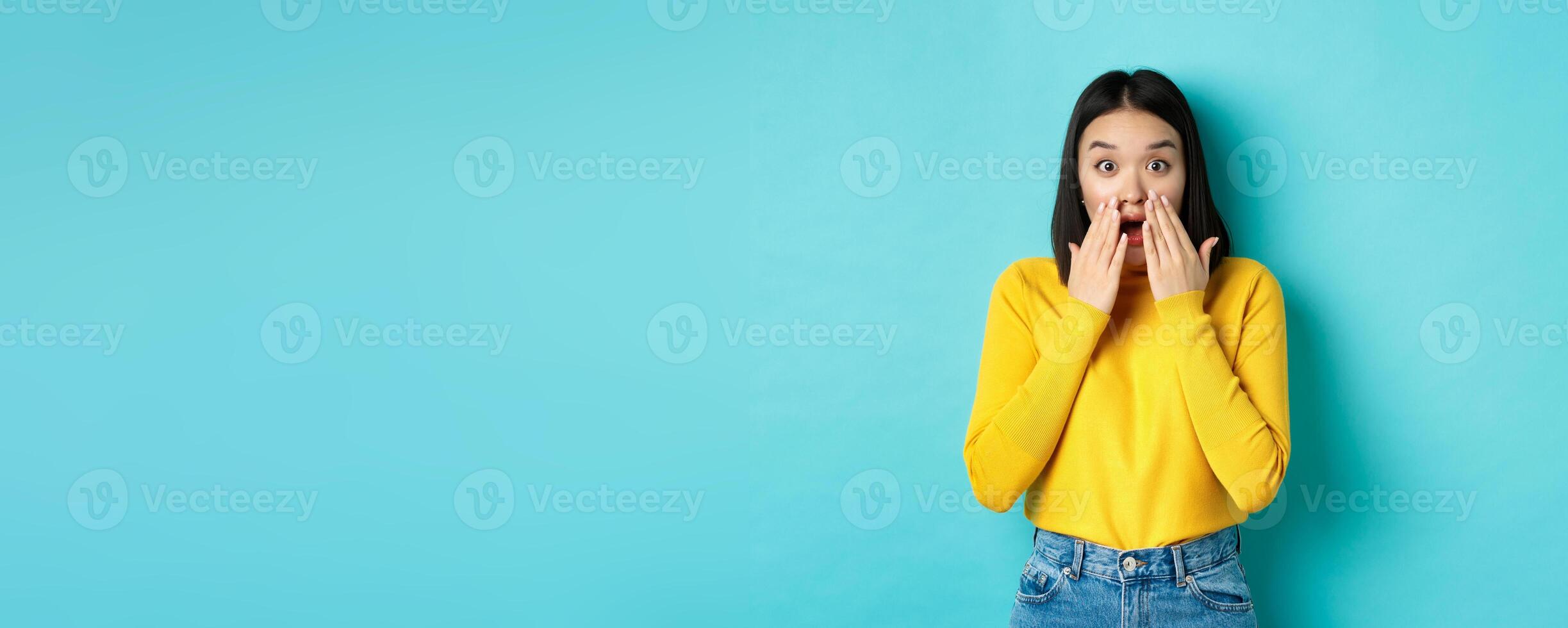 Portrait of impressed korean girl saying wow, gasping and staring amazed at camera, standing in yellow pullover over blue background photo