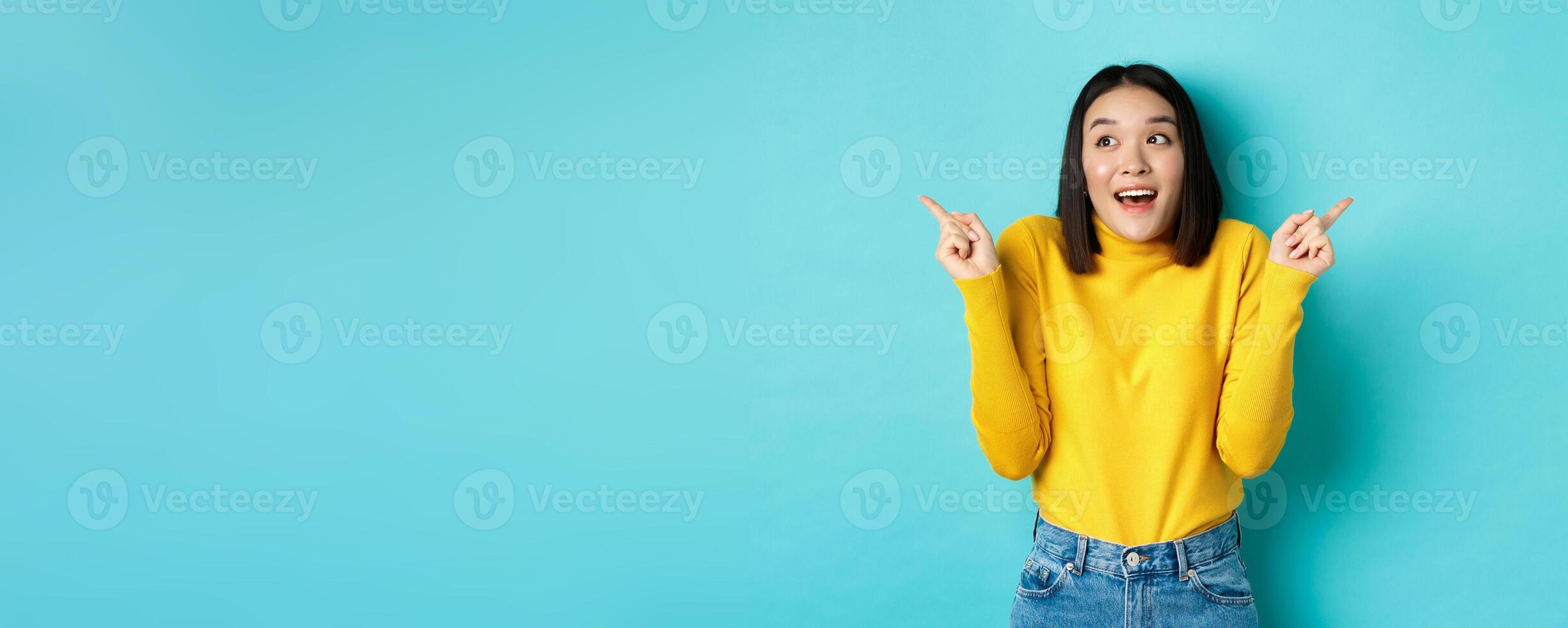 Beauty and fashion concept. Excited asian girl in yellow pullover making choice, pointing fingers sideways and choosing between two options, standing against blue background photo
