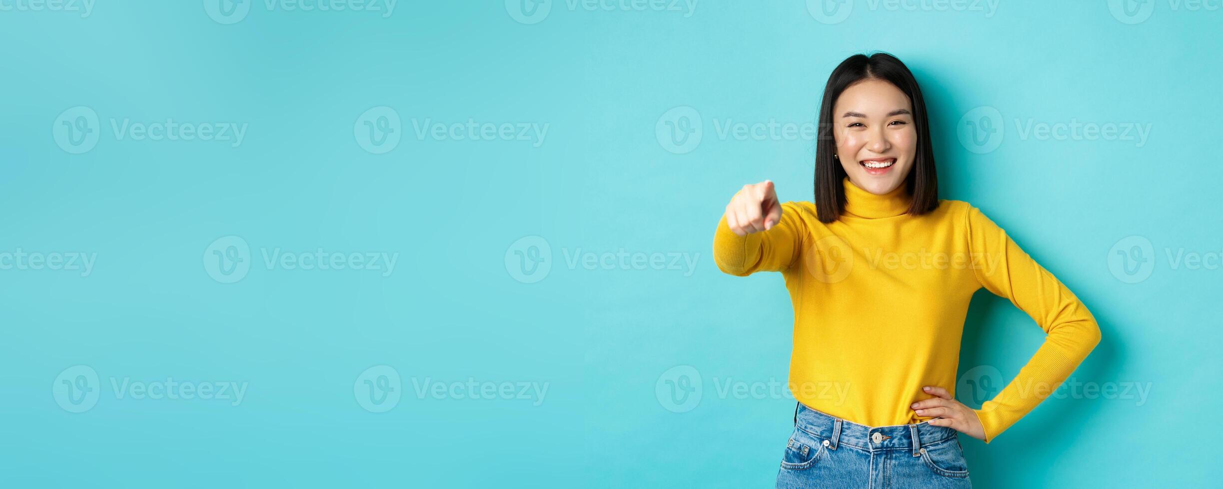 Beauty and fashion concept. Cheerful asian woman laughing and smiling, pointing finger at camera, choose you, standing over blue background photo