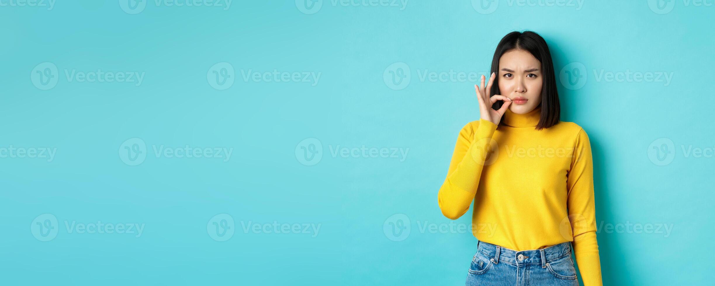 Serious asian girl showing mouth zip gesture, promise to keep shut and frowning, telling big secret, standing in yellow pullover over blue background photo
