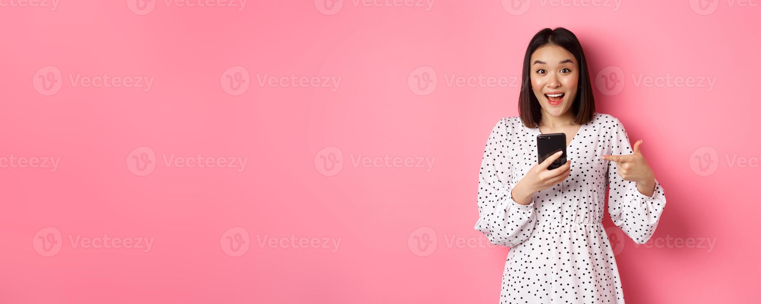 Online shopping and beauty concept. Amazed and happy asian woman pointing at mobile phone, talking about internet promo offer or app, standing over pink background photo