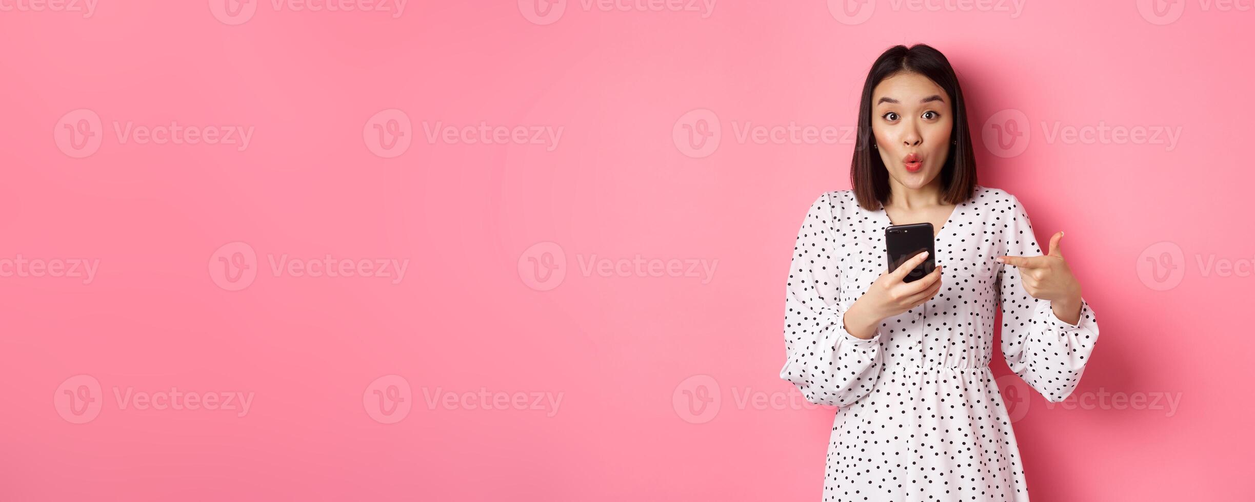 Online shopping and beauty concept. Amazed asian woman pointing finger at mobile phone, recommending smartphone app, standing over pink background photo