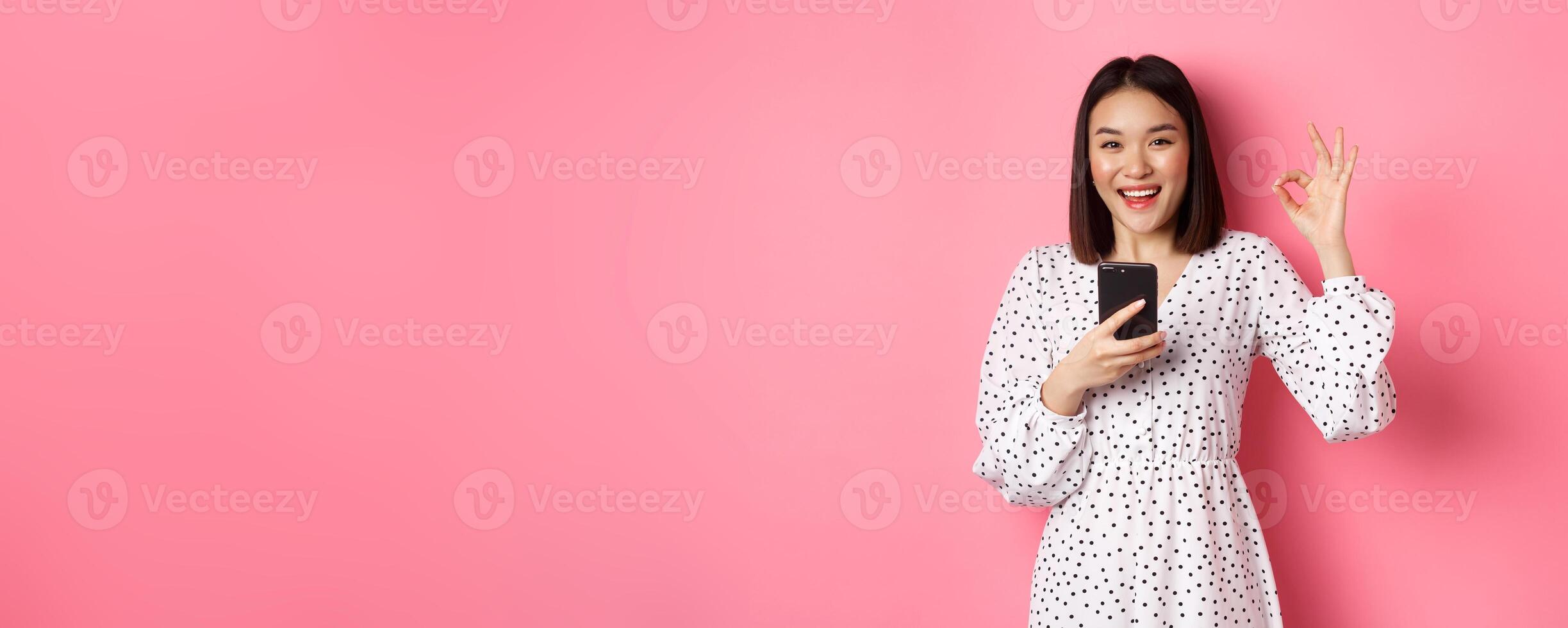 Online shopping and beauty concept. Satisfied asian female customer showing okay, making purchase in internet on smartphone, standing over pink background photo