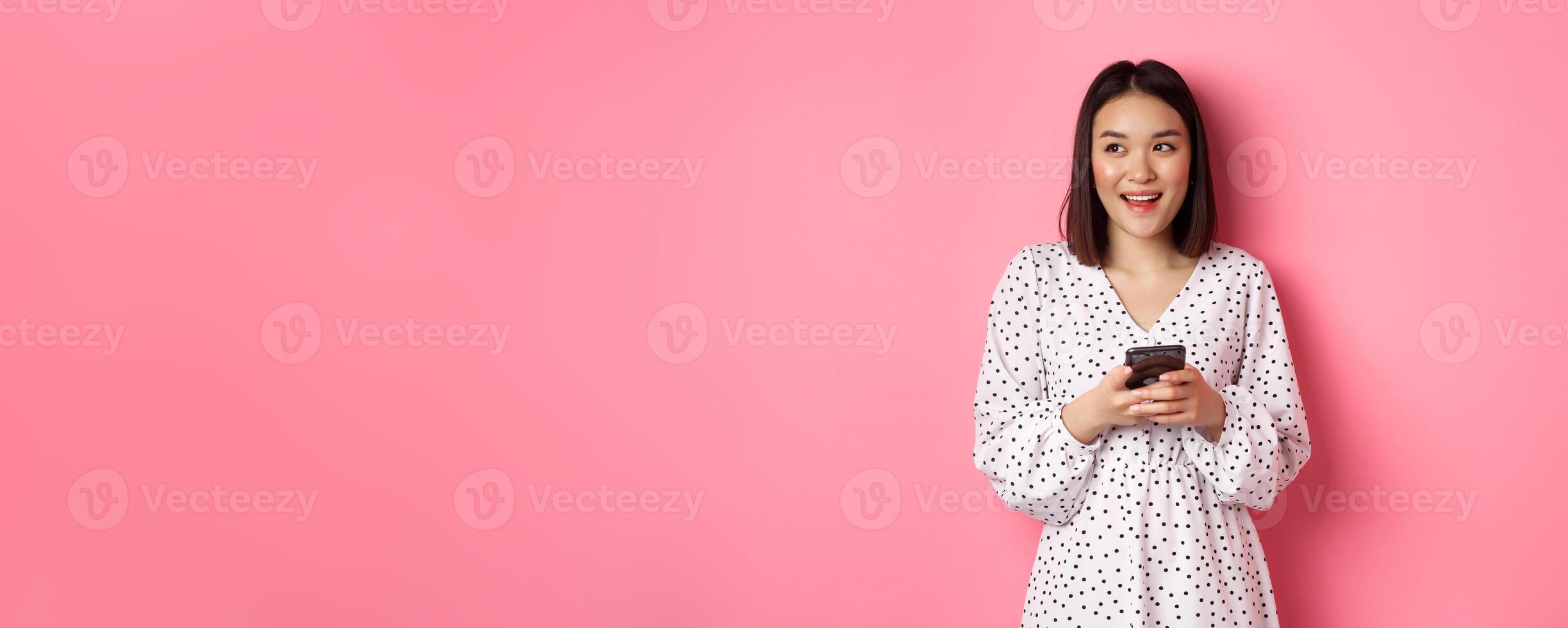 Cute asian woman thinking and smiling, looking left dreamy while messaging on smartphone, browing online stores, standing over pink background photo