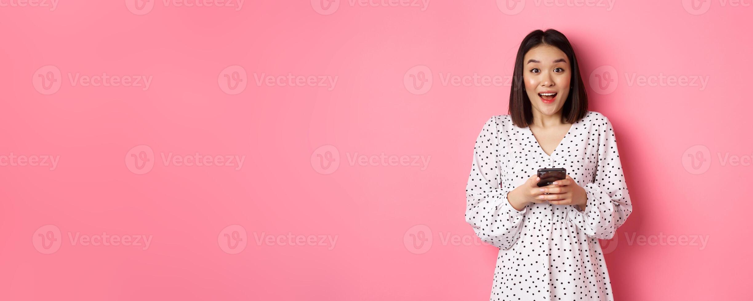 Online shopping. Amazed asian woman looking at camera with happy smile, making purchase with smartphone, using mobile phone app, standing over pink background photo