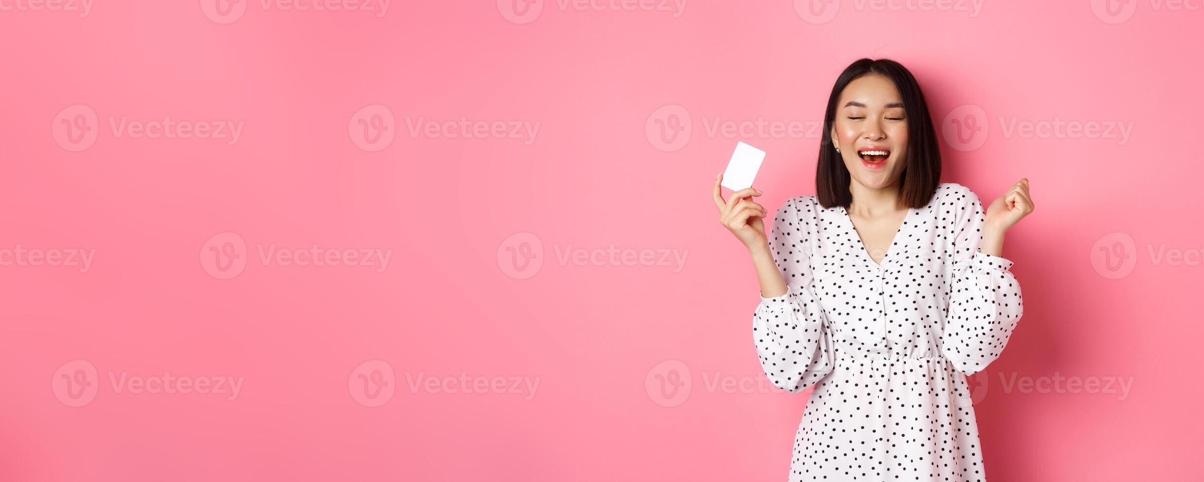 Shopping concept. Excited asian woman feeling satisfaction, holding credit card and rejoicing, standing over pink background photo