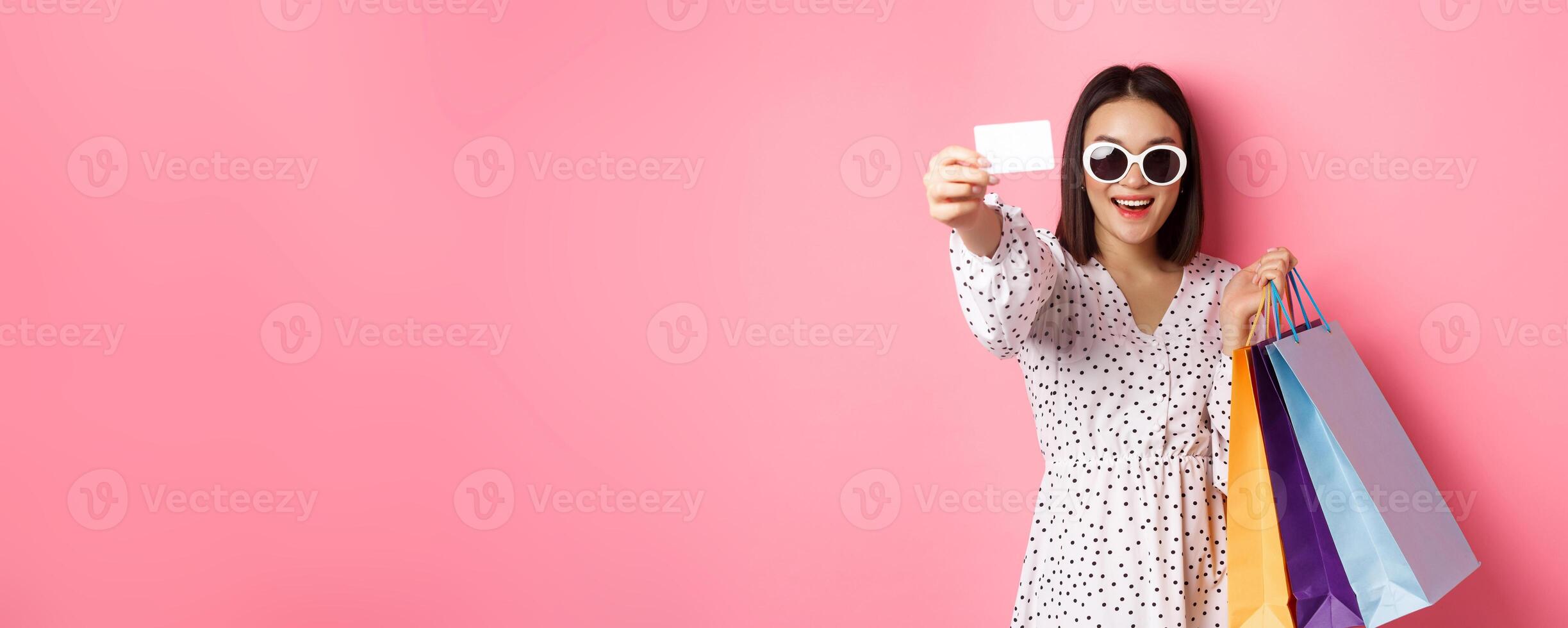 Beautiful asian woman in sunglasses going shopping, holding bags and showing credit card, standing over pink background photo