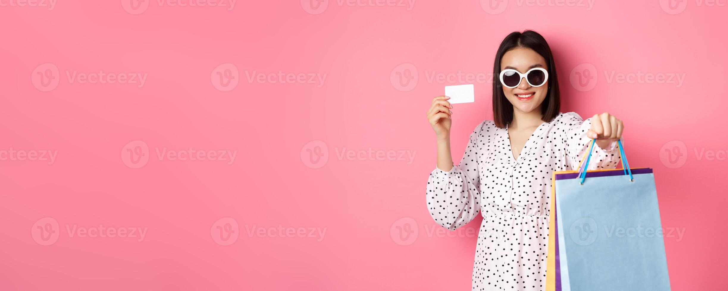 Beautiful asian woman in sunglasses going shopping, holding bags and showing credit card, standing over pink background photo