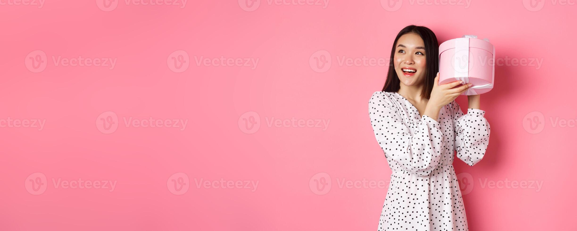 Cute asian woman shaking box with gift, smiling and looking intrigued, guess what inside present, standing over pink background photo