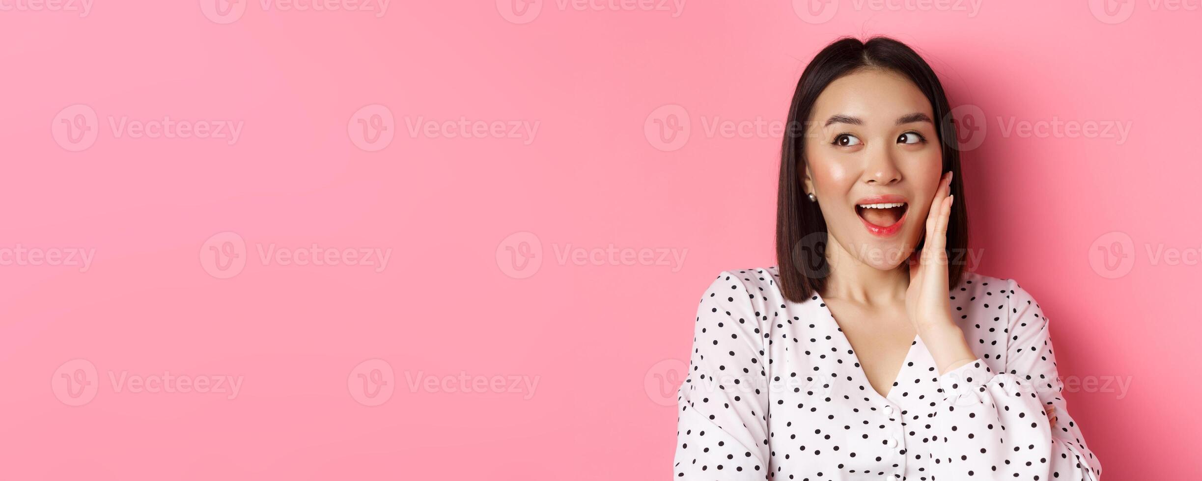 Coquettish asian woman in blushing, touching cheek and looking left amused, standing over pink background photo