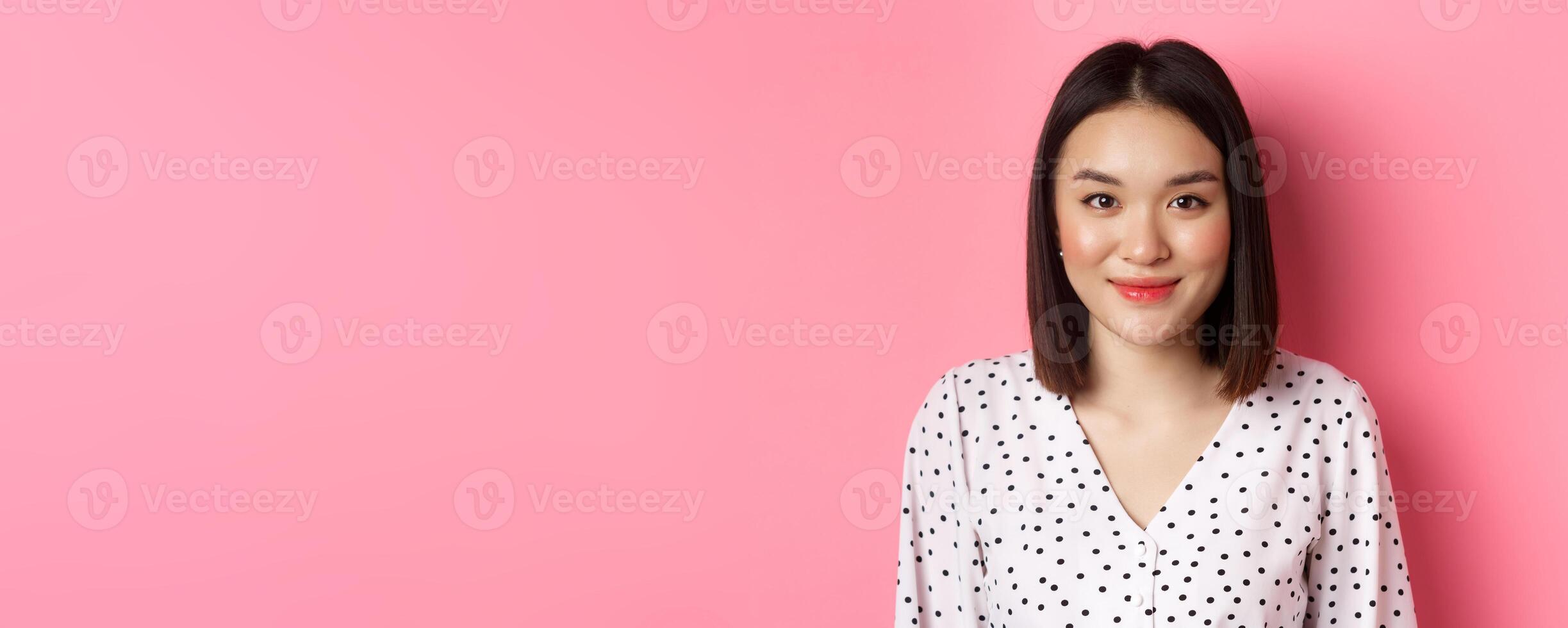 Close-up of cute asian female model looking at camera, blushing and smiling kawaii, standing over pink background photo