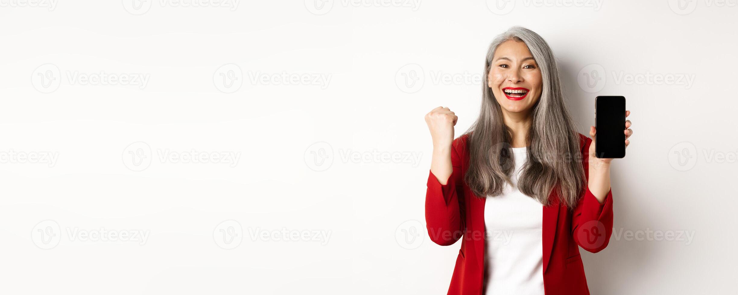 Success. Asian senior woman showing smartphone blank screen and fist pump, winning prize online, standing over white background photo