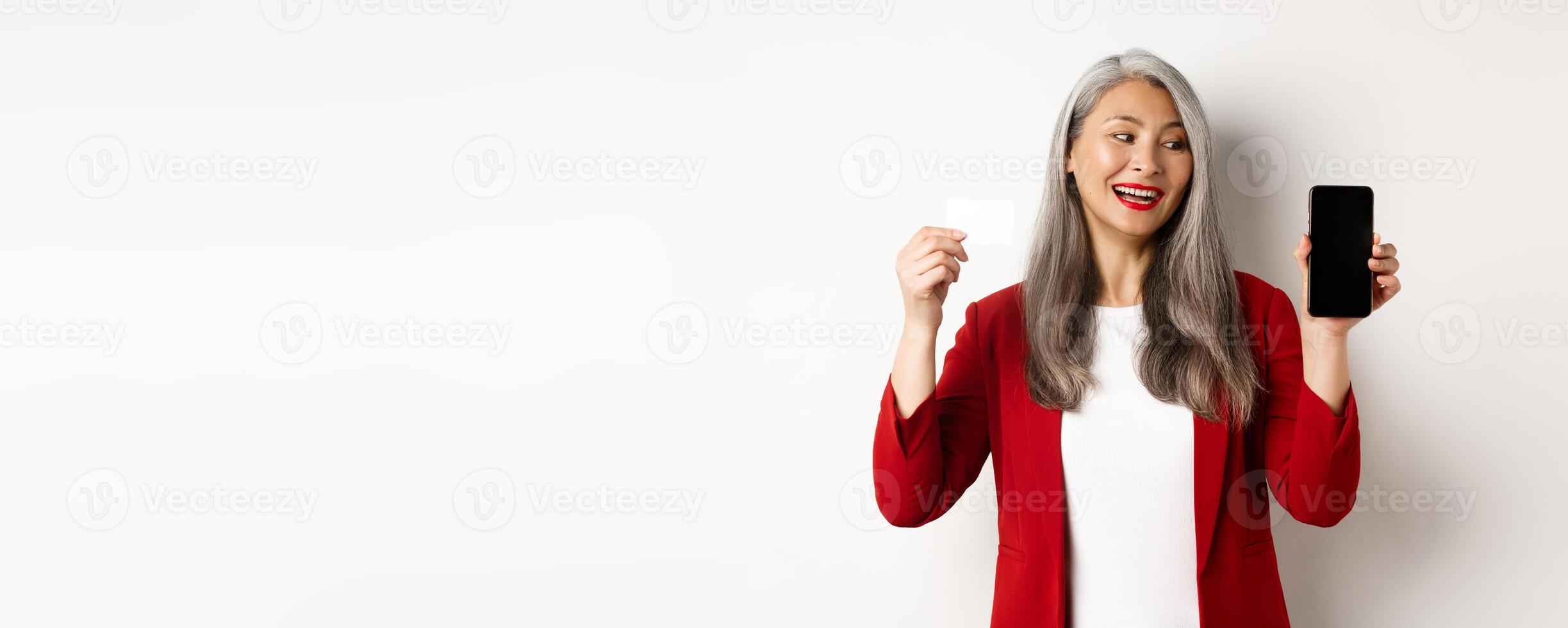 Cheerful asian mature woman showing blank smartphone screen and credit card, concept of e-commerce photo