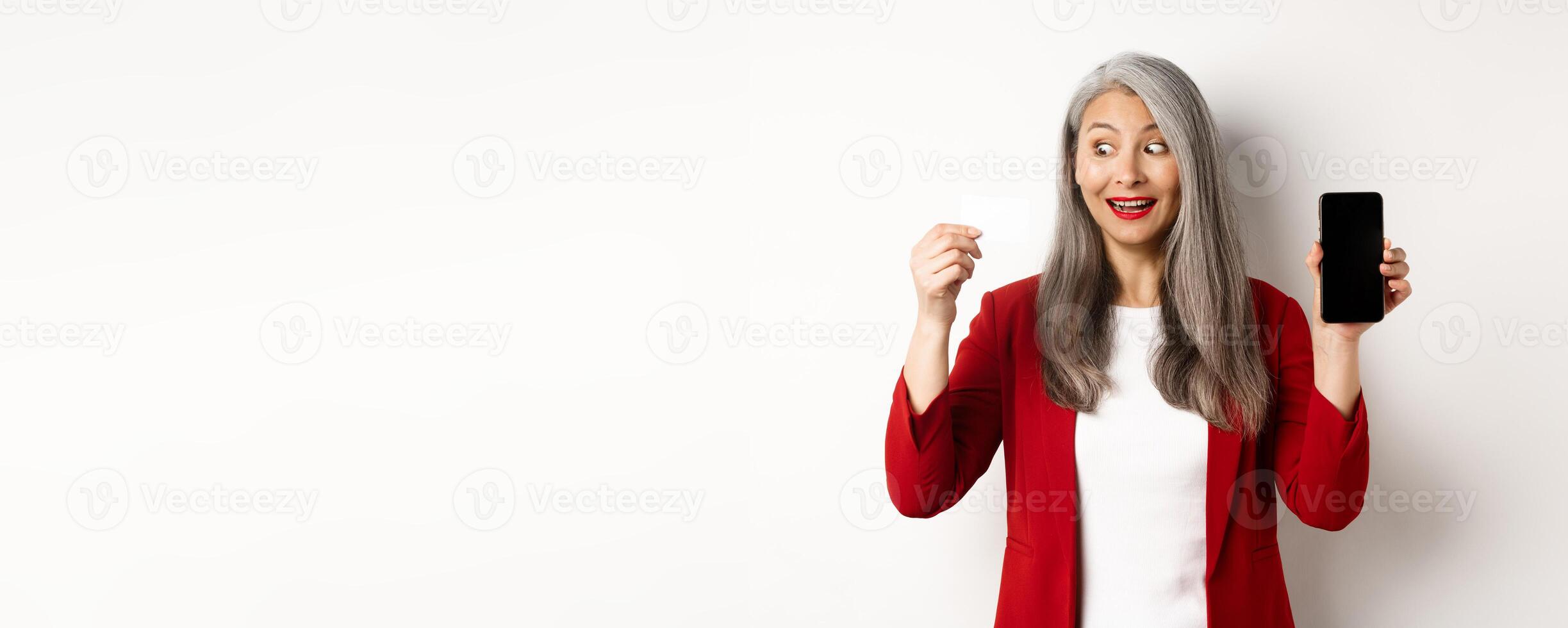 Cheerful asian mature woman showing blank smartphone screen and credit card, concept of e-commerce photo